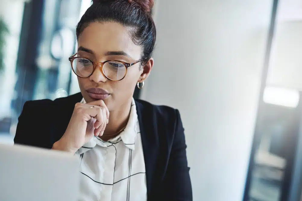 Woman in glasses thinking at her office laptop, weighing a decision